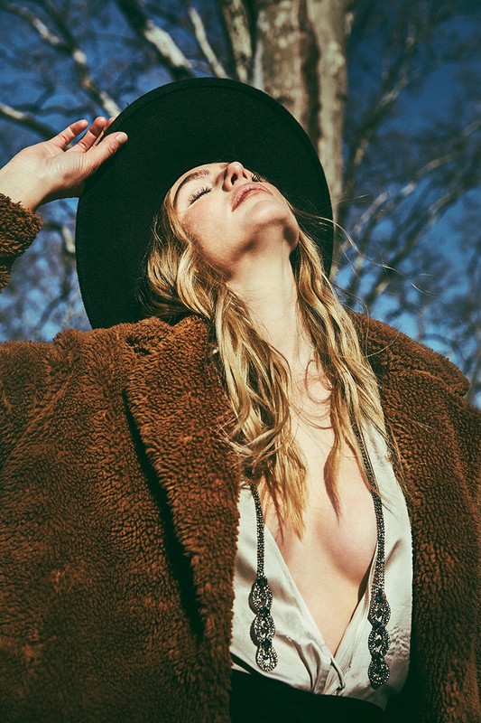 Stylish woman in brown teddy coat and black hat poses outdoors with a tree branch and blue sky in the background during daylight.