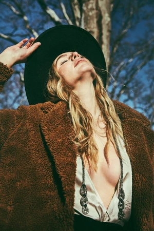 Stylish woman in brown teddy coat and black hat poses outdoors with a tree branch and blue sky in the background during daylight.