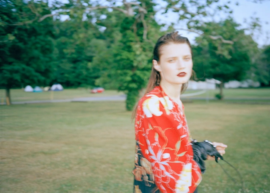 A woman in a floral top stands in a grassy park with trees and tents in the background in a medium shot with a shallow depth of field.