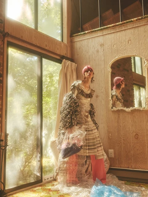 Indoor fashion portrait of a woman in a transparent outfit with bubble wrap, standing in front of a large window in a wood-paneled room.