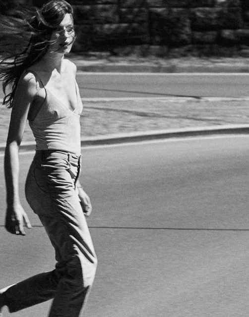 Monochrome shot of a woman with long brown hair walking on an urban street wearing a tank top and pants on a sunny day.