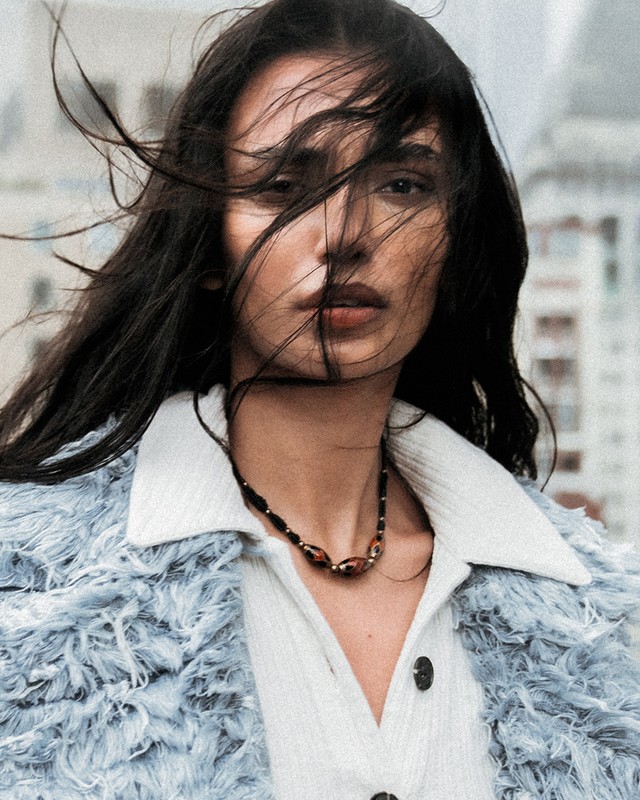 Close-up portrait of a woman with dark hair blowing in the wind, wearing a light blue jacket, against an urban background.