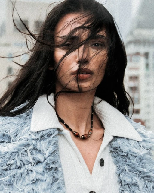 Close-up portrait of a woman with dark hair blowing in the wind, wearing a light blue jacket, against an urban background.