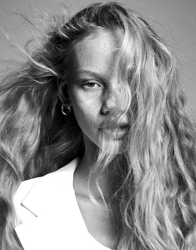 Close-up monochrome portrait of a woman with long wavy blonde hair blowing across her face and wearing a white blazer.