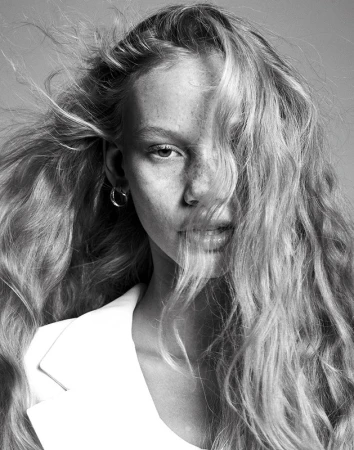 Close-up monochrome portrait of a woman with long wavy blonde hair blowing across her face and wearing a white blazer.
