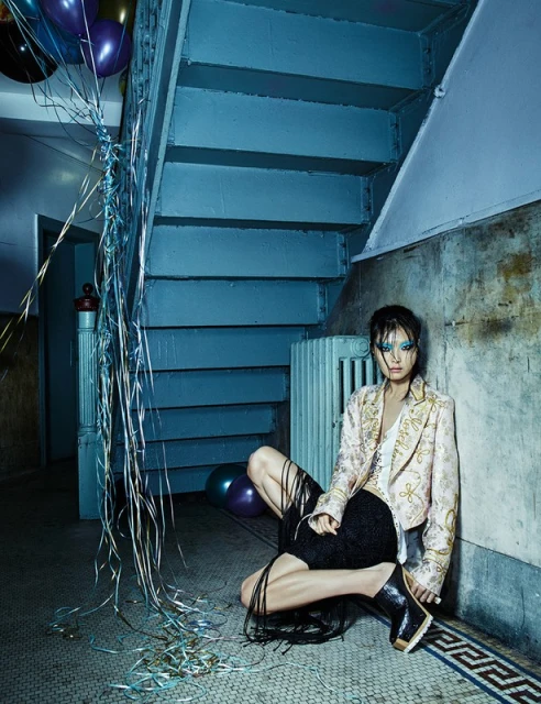 A female model poses in an indoor staircase area near silver streamers, wearing a pale gold blazer, black shorts, and shiny black platform boots.