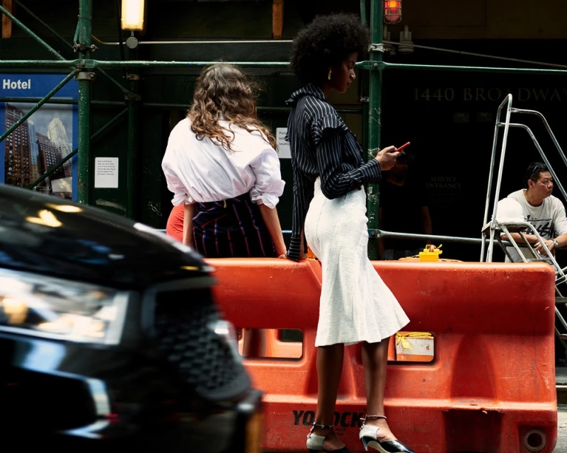 Two women in stylish outfits are on a city street near a black car, with one woman looking at her phone, captured in daylight.