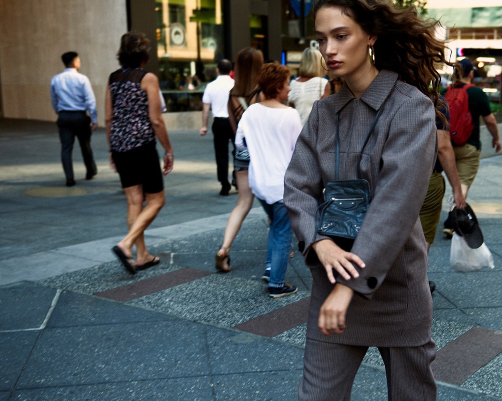 Woman in corduroy suit stands out in city crowd, carrying a small bag, accessorized with hoop earrings, urban backdrop, candid.