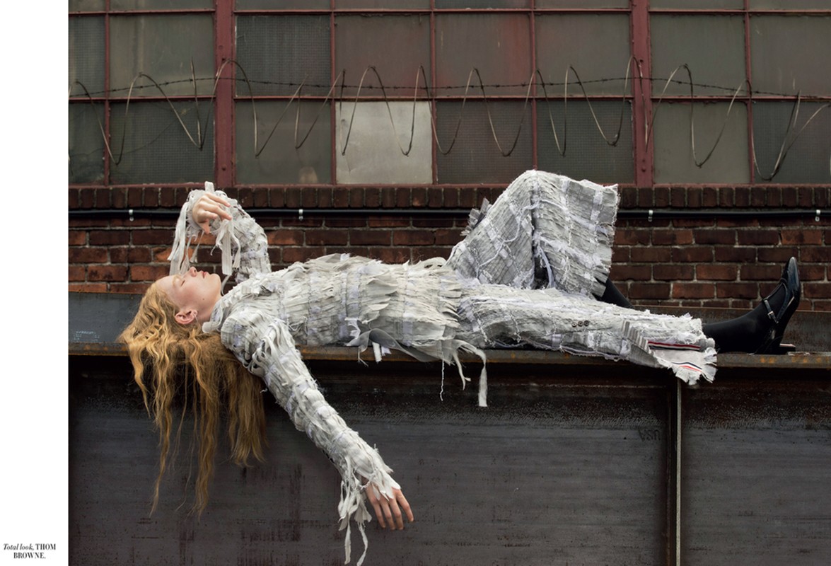 Woman in a textured white outfit lounges on a weathered gray ledge in front of a red brick wall with barred windows and barbed wire.