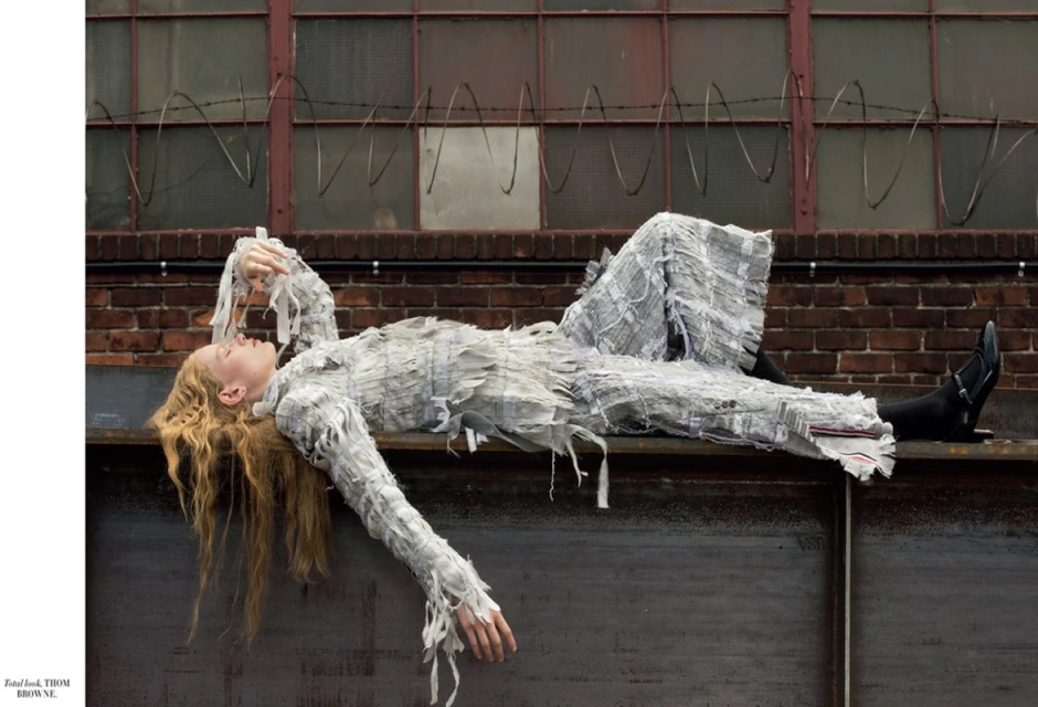 Woman in a textured white outfit lounges on a weathered gray ledge in front of a red brick wall with barred windows and barbed wire.