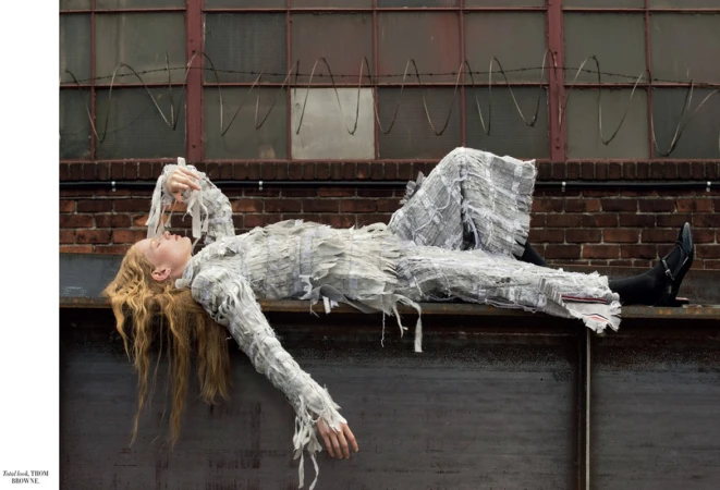 Woman in a textured white outfit lounges on a weathered gray ledge in front of a red brick wall with barred windows and barbed wire.
