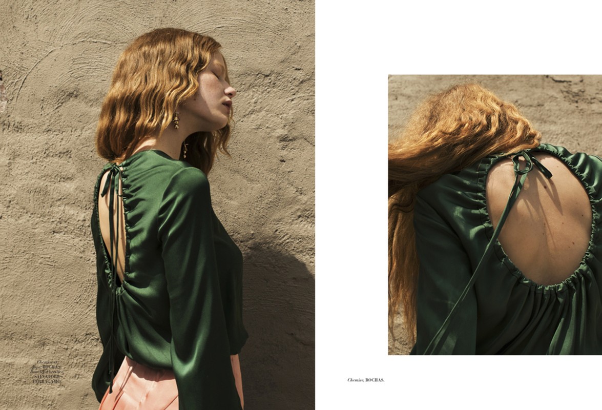 Fashion shot of a woman with auburn hair wearing a green top and pink skirt, standing in front of a textured wall in natural light.