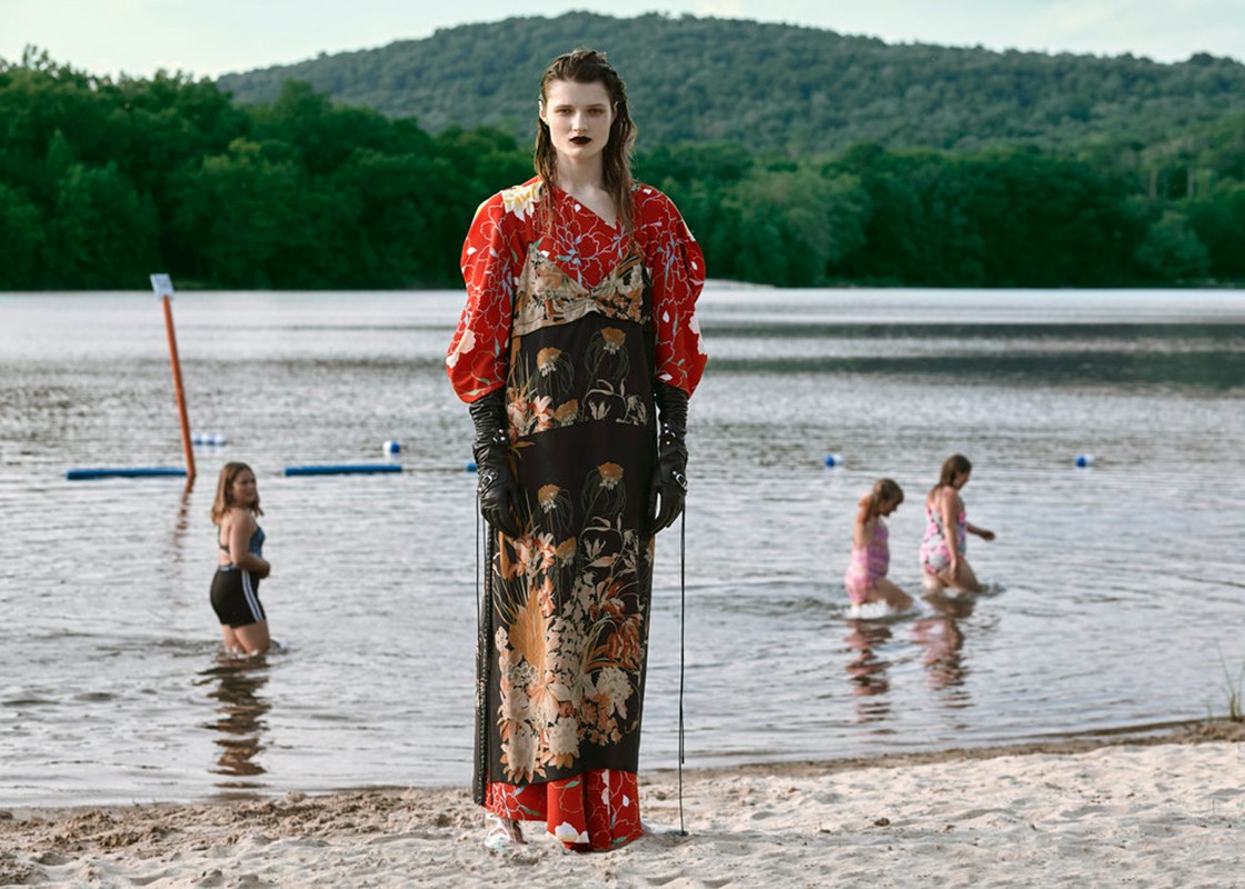 A fashionable adult female model in a colorful gown stands on a sandy beach near a lake with children wading in the water and trees visible.