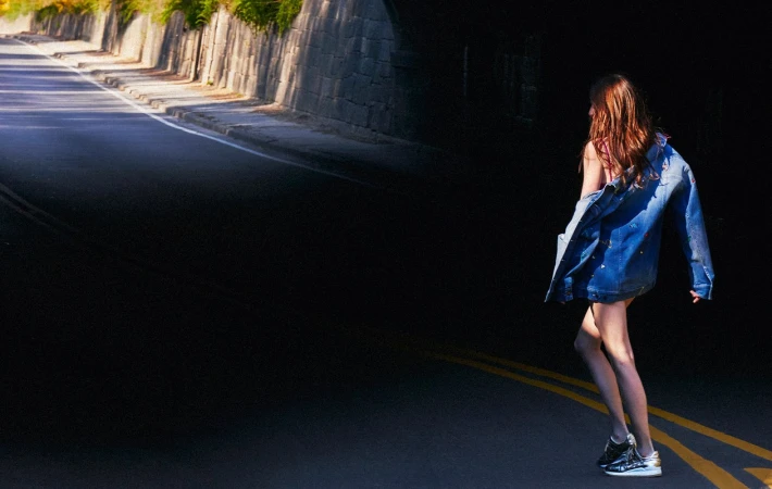A woman in a denim jacket walks on an asphalt road leading into a dark tunnel with a stone wall and green vegetation on top.