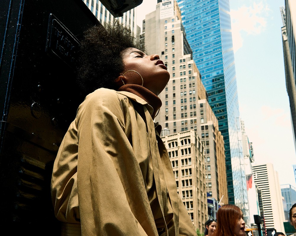 Fashionable woman with afro in trench coat gazes upward in a city, near a black metal structure, surrounded by tall buildings on a sunny day.