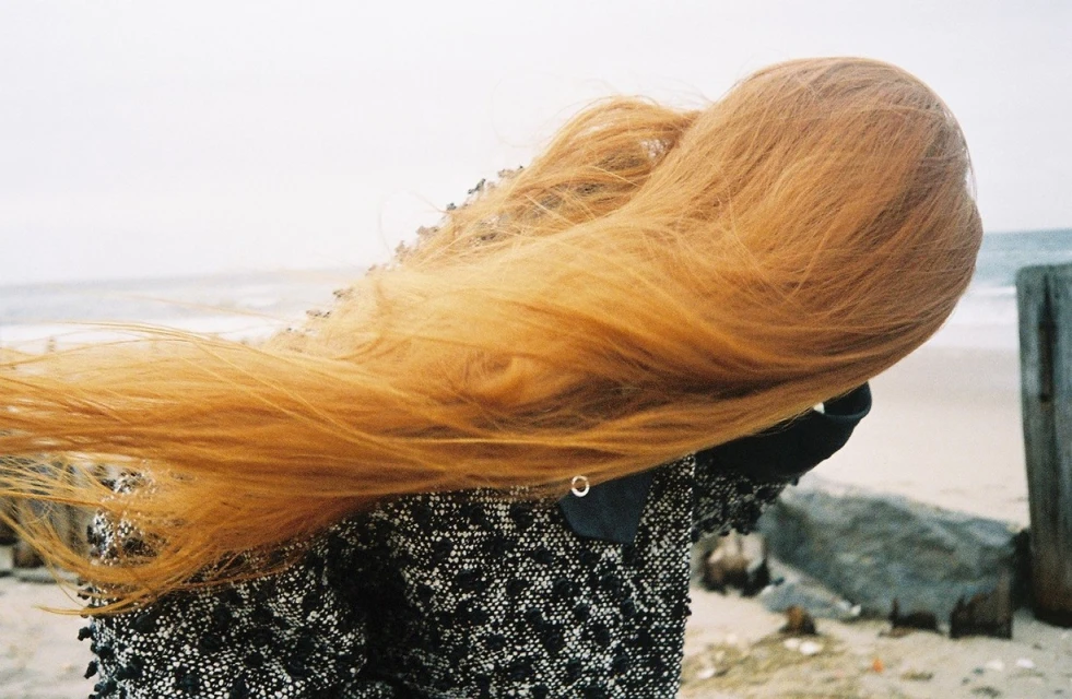 Woman with flowing red hair poses on a beach in a tweed jacket on an overcast day with the ocean and sky blurred in the background.