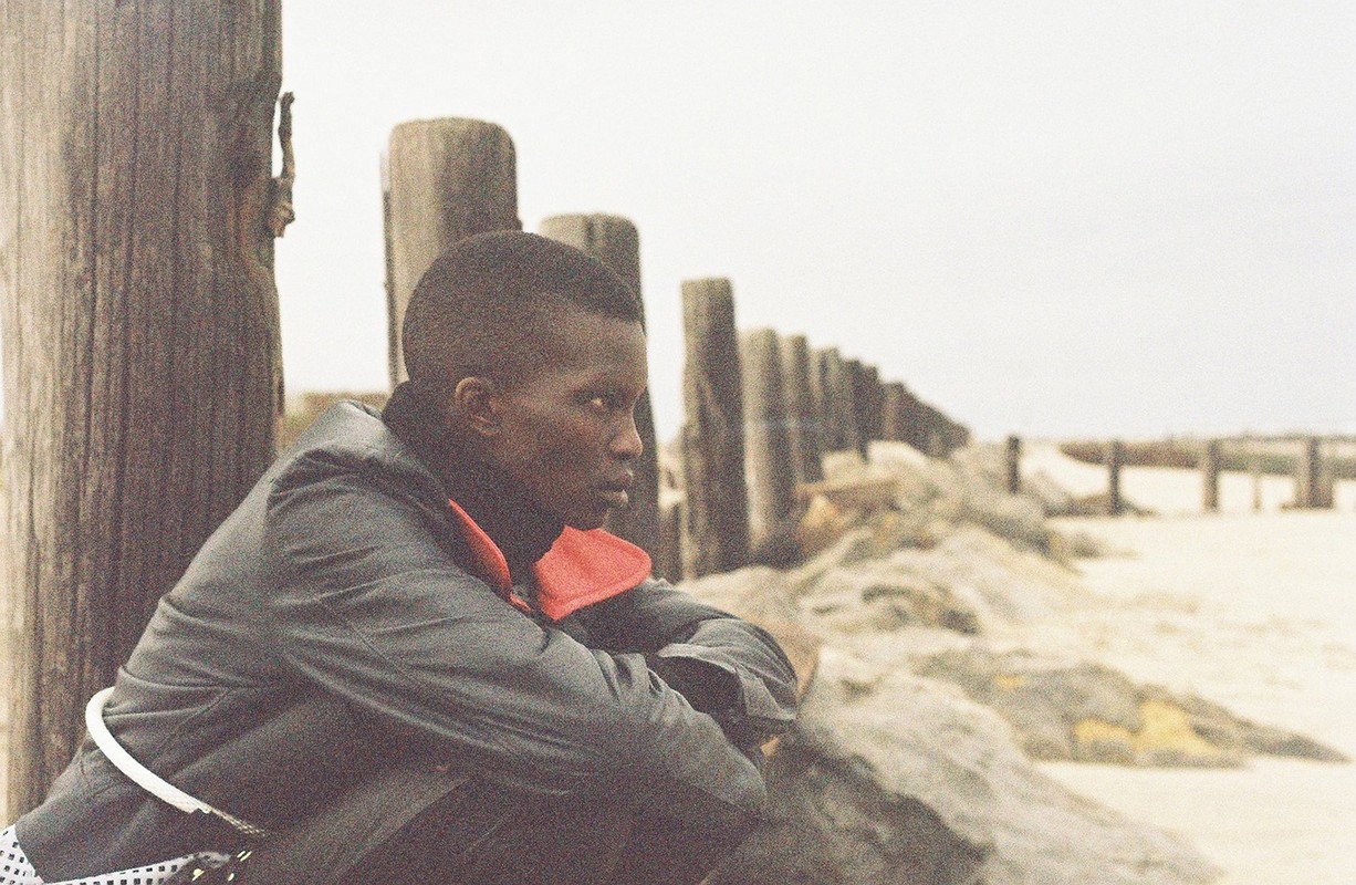 An adult sits beside a row of wooden posts along a sandy beach under an overcast sky, looking out towards the horizon in contemplation.