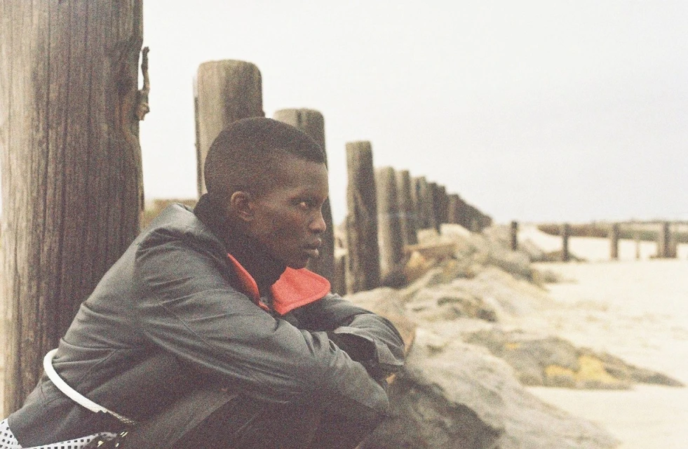 An adult sits beside a row of wooden posts along a sandy beach under an overcast sky, looking out towards the horizon in contemplation.