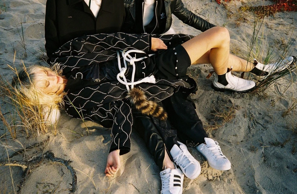 Eye-level shot on sand of two people wearing black and white clothing and Adidas sneakers, one lying down and one sitting.