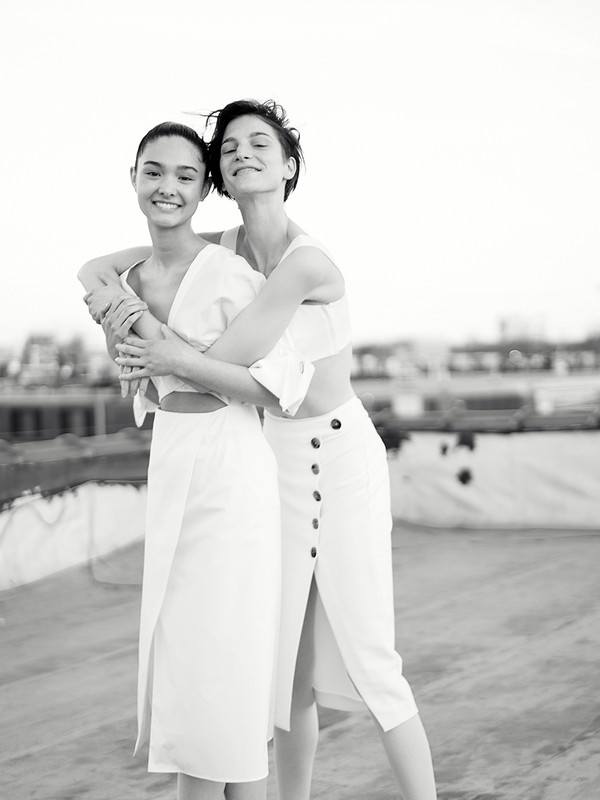 Two women in white dresses embrace on a rooftop with a cityscape backdrop. They exude a natural, candid mood, capturing a moment of connection.