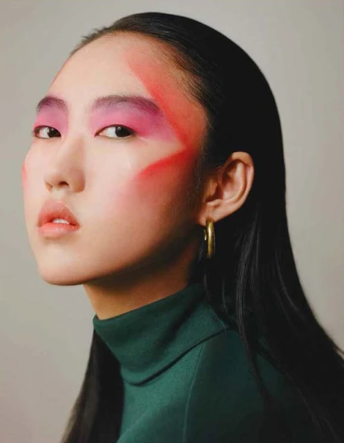 Close-up of Asian woman with dramatic pink eye makeup and green turtleneck, showcasing beauty and fashion in a minimalist studio.