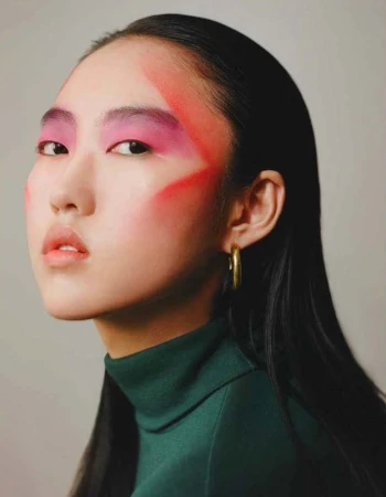 Close-up of Asian woman with dramatic pink eye makeup and green turtleneck, showcasing beauty and fashion in a minimalist studio.