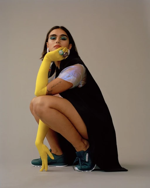 Woman in a black dress and yellow gloves squats wearing green sneakers against a grey background in a fashion studio shot.