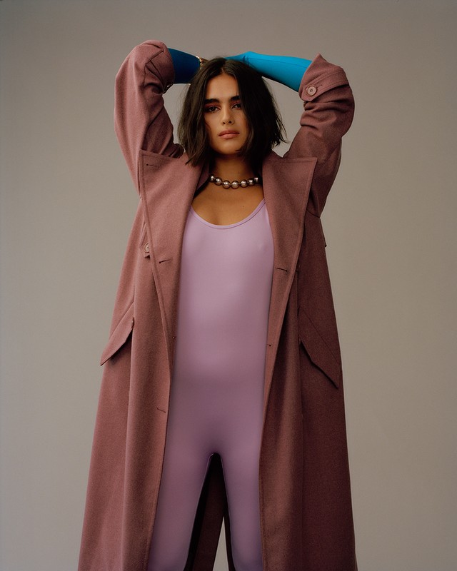 A woman poses indoors wearing a lilac catsuit, blue gloves, a large pearl necklace, and a dusty-rose trench coat.
