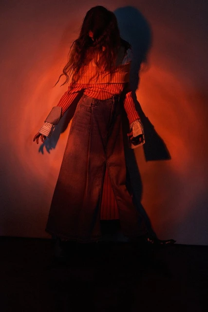 Fashion photo of a person in a long denim skirt, striped top, and long hair standing in front of a red-lit wall, creating a dramatic shadow.