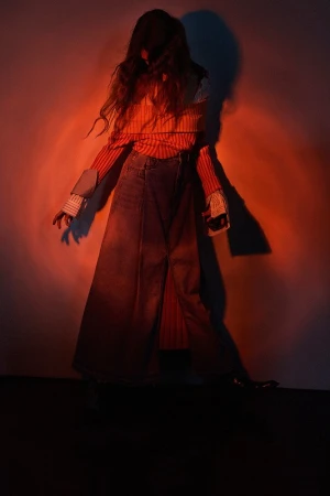 Fashion photo of a person in a long denim skirt, striped top, and long hair standing in front of a red-lit wall, creating a dramatic shadow.
