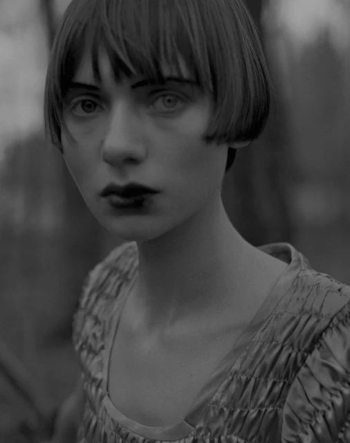 Monochrome portrait of a young woman with dark makeup, bob haircut, and a silky dress, posing outdoors in a blurred forest-like setting.