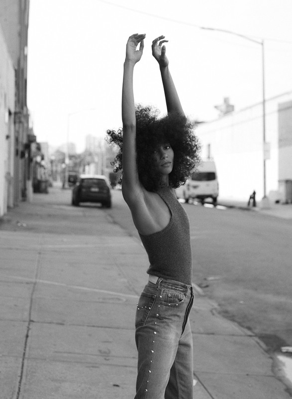 Monochrome portrait of a woman with an afro hairstyle posing on a city street in a tank top and embellished jeans with arms raised.