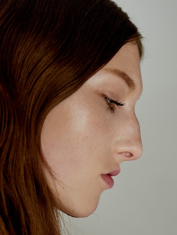 Close-up profile portrait of a woman with long brown hair and natural makeup against a plain light background.