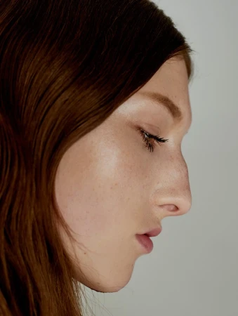 Close-up profile portrait of a woman with long brown hair and natural makeup against a plain light background.