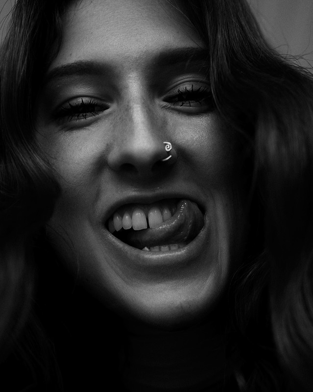 Black and white portrait of a young woman sticking out her tongue, wearing a nose ring with curly hair. 