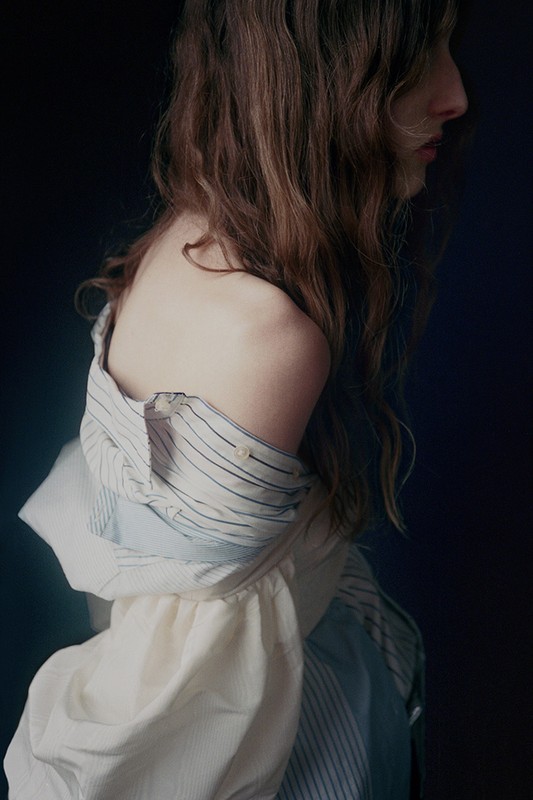 Profile portrait of a woman with long wavy brown hair wearing an off-the-shoulder striped shirt against a dark background.
