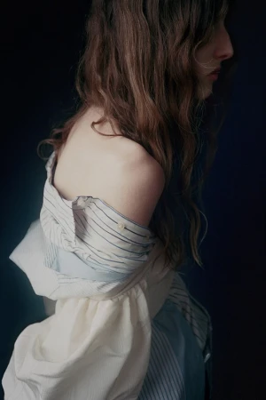 Profile portrait of a woman with long wavy brown hair wearing an off-the-shoulder striped shirt against a dark background.