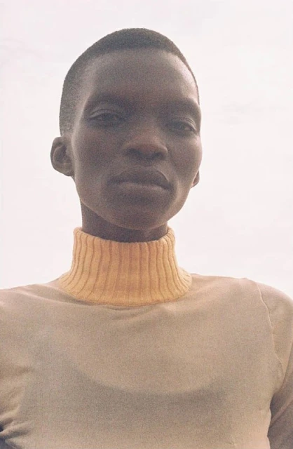 Headshot of an African female in a beige turtleneck. The lighting is soft, and she looks directly into the camera.