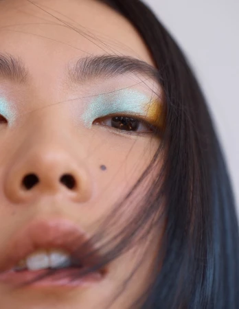 Close-up portrait of an Asian adult female face with creative eye makeup and dark hair, against a plain light backdrop in indoor setting.