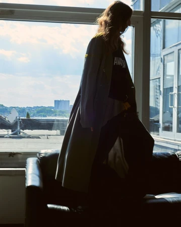 Model wearing a grey trench coat and dark clothing standing on a sofa near a large window overlooking an outdoor view.