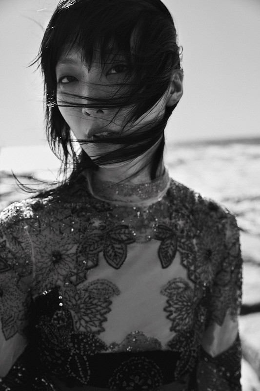 Monochrome portrait of an Asian female model with dark hair, gazing at the camera outdoors with a high-necked floral embellished garment.