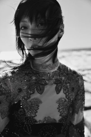 Monochrome portrait of an Asian female model with dark hair, gazing at the camera outdoors with a high-necked floral embellished garment.