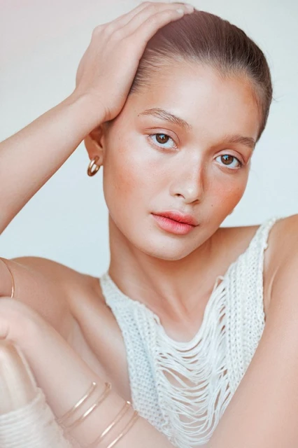 Close-up portrait of a young adult female model with hand on head, wearing a white top and gold hoop earrings in a light setting.