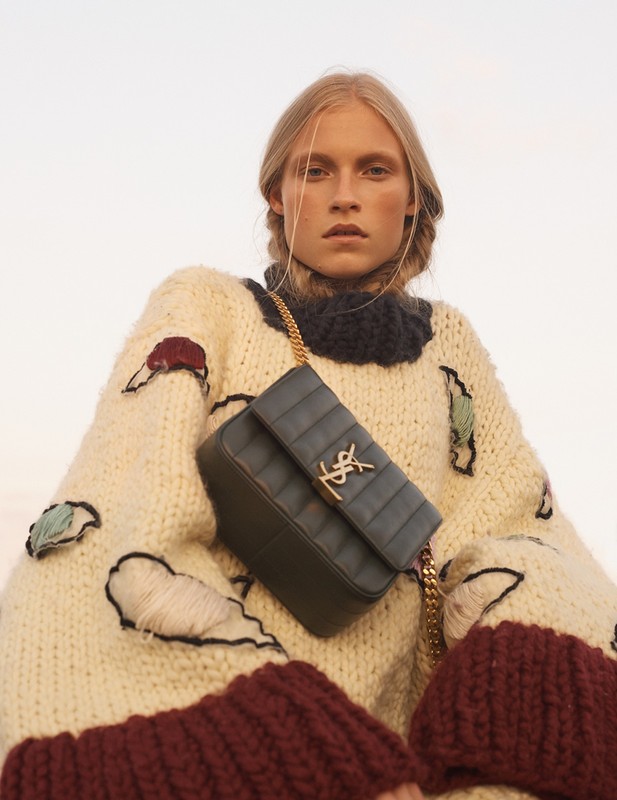 Blonde woman in knit sweater with blue leather YSL bag, posing outdoors in warm light for a fashion editorial. Minimalist background.
