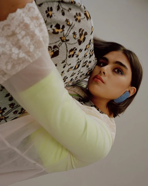 Portrait of a young woman with bobbed brown hair and blue earrings wearing a yellow top and sheer white blouse with floral accents.