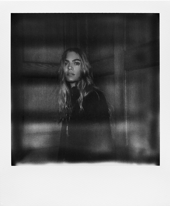 Portrait of a young adult woman with long blond hair and dark clothing, in a soft-focus black and white photograph against a dark background.