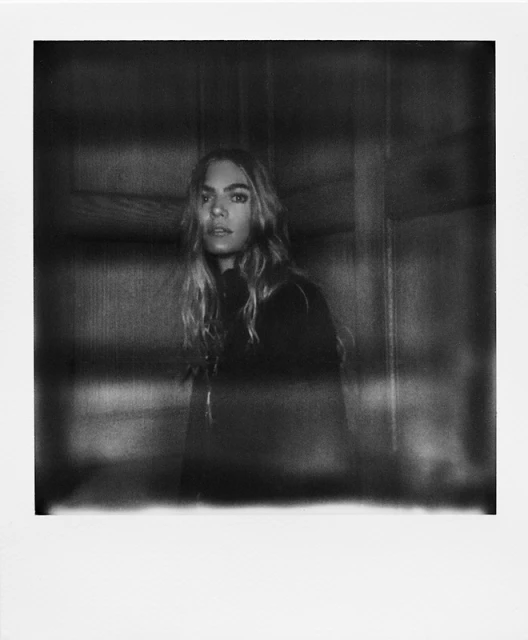 Portrait of a young adult woman with long blond hair and dark clothing, in a soft-focus black and white photograph against a dark background.