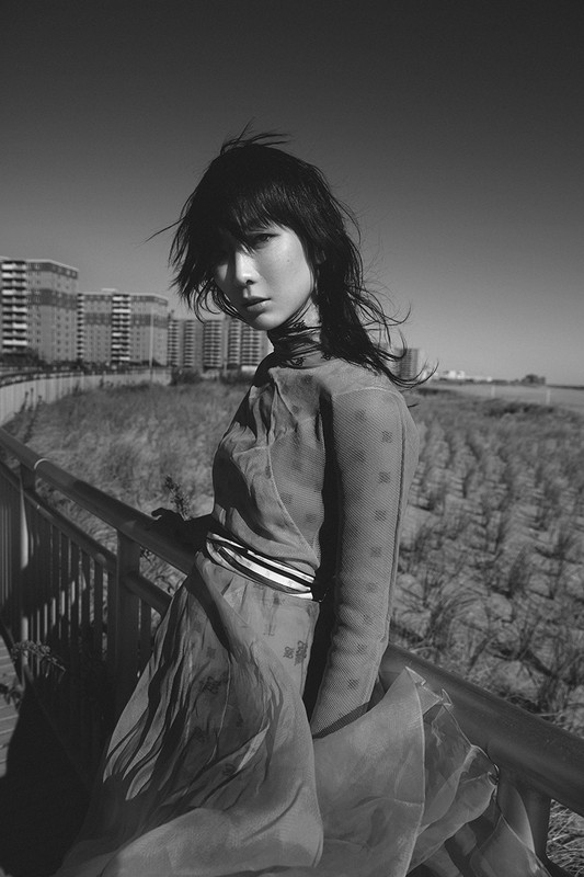 Black and white portrait of a young woman in a flowing dress, standing on a boardwalk with buildings and sky in the background.