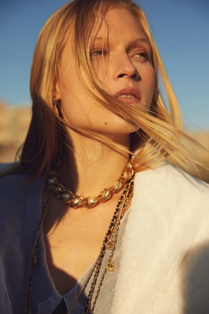 Close up portrait of a blonde woman wearing gold chain necklaces and a white textured garment outdoors at golden hour.