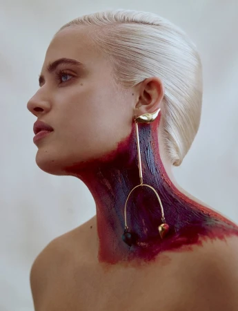 Profile portrait of a blonde woman wearing a large gold drop earring, with red and purple paint smeared on her neck